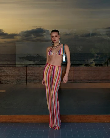 Fashionable adult woman poses in a multicolored knit bikini and pants with a modern handbag against a sunset ocean view.