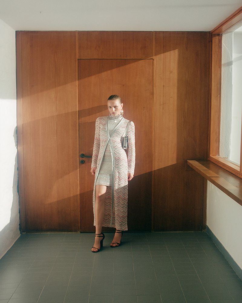 A stylish adult woman in a light chevron-pattern coat and mini dress poses indoors in front of a wooden wall and door, accessorized with sandals.