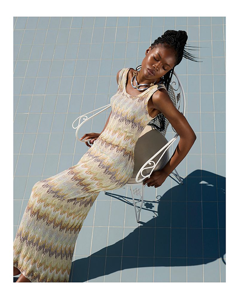 Woman in patterned maxi dress and necklace sits in white chair on light blue tile in bright sunlight casting strong shadows. 