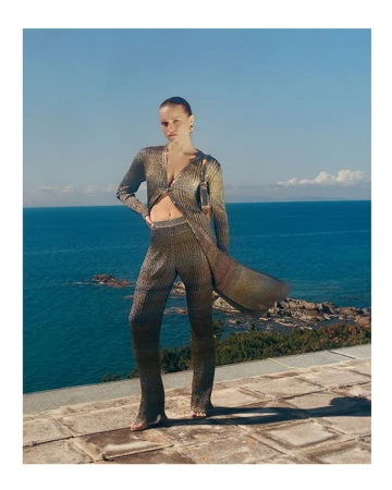 A woman poses in a sparkly gold knit set on a stone patio with an ocean view, wearing a matching shoulder bag in natural light.