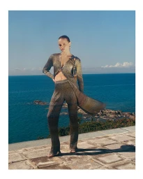 Model wearing a shimmering metallic gold knit cardigan and matching trousers standing outdoors against an ocean view backdrop.