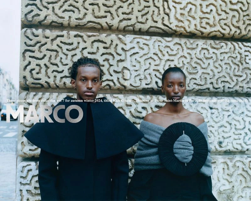 Two individuals in avant-garde fashion pose outdoors against an textured stone wall on a city street for a fashion editorial shoot.