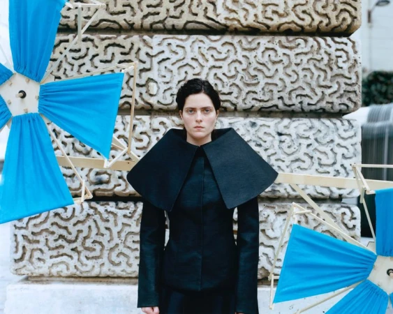 Adult woman in black wide-collar jacket poses outdoors near stacked concrete architectural blocks and blue fabric pinwheel art installation.
