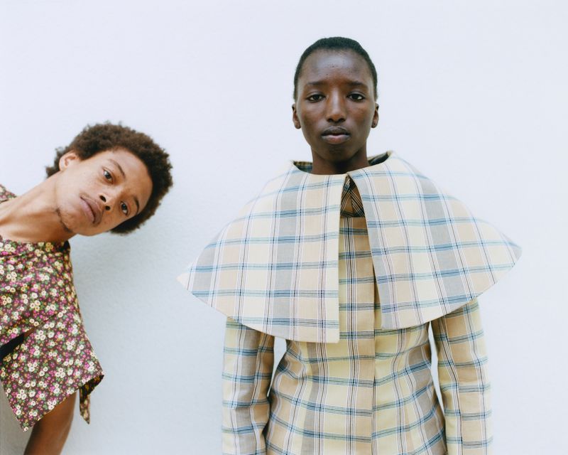 Two people in a studio setting. One person in a plaid outfit and another in a floral top. Minimalistic fashion portrait with clean backdrop.
