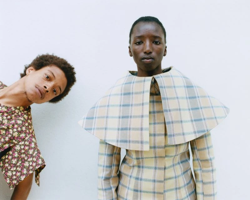 Two people in a studio setting. One person in a plaid outfit and another in a floral top. Minimalistic fashion portrait with clean backdrop.