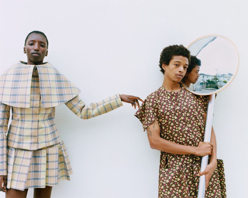 Two people in vintage-style clothing stand in front of a white wall. One model holds a round mirror that reflects a cityscape.