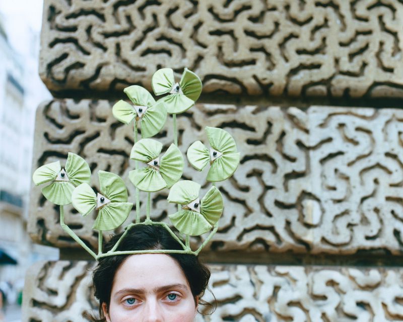 Model with creative light green floral headpiece stands before a carved stone wall in an outdoor urban environment. Fashion editorial.