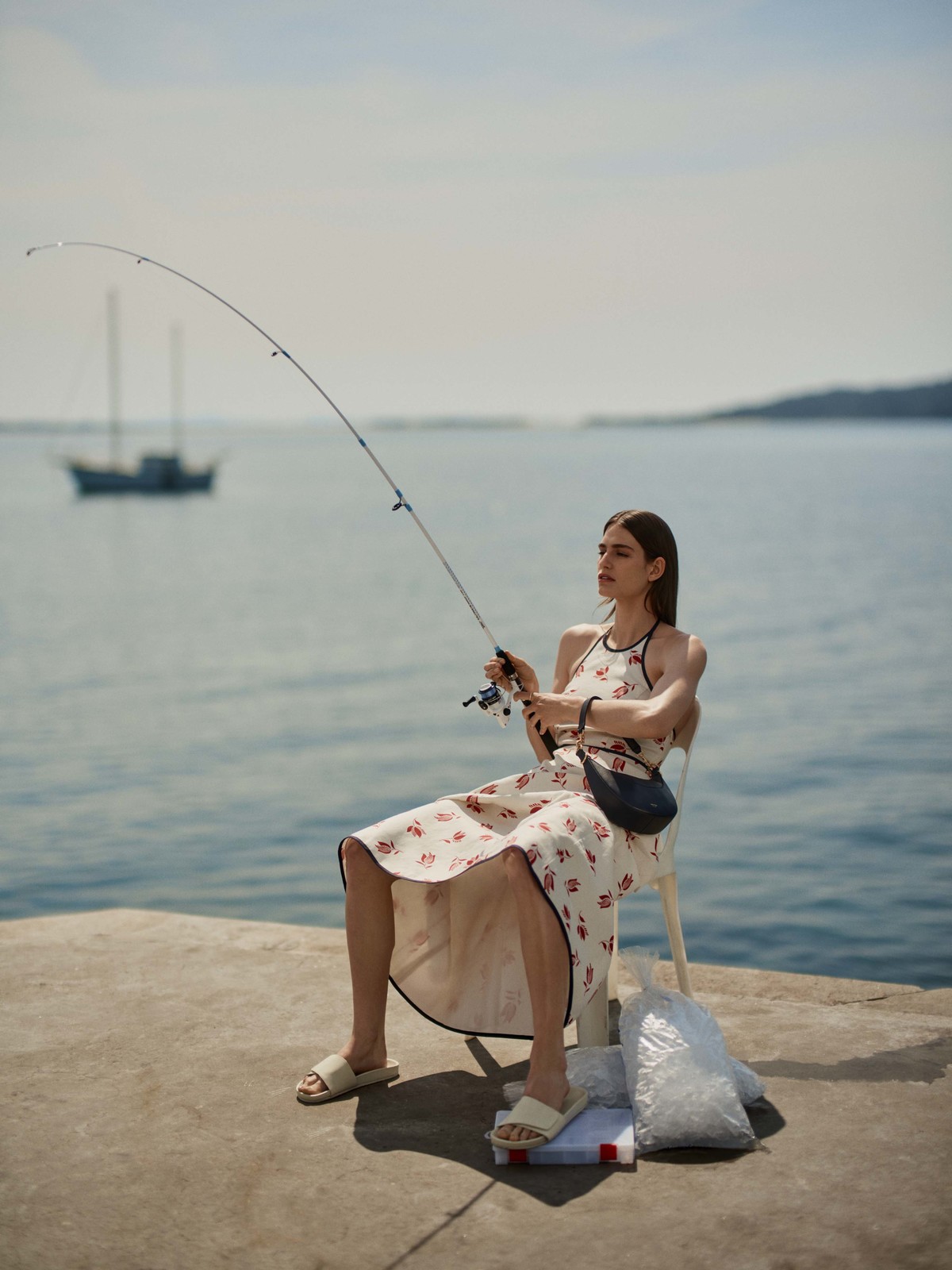 A woman in a floral dress sits on a chair, fishing rod in hand, at the water's edge, with a boat in the background and blue sky.