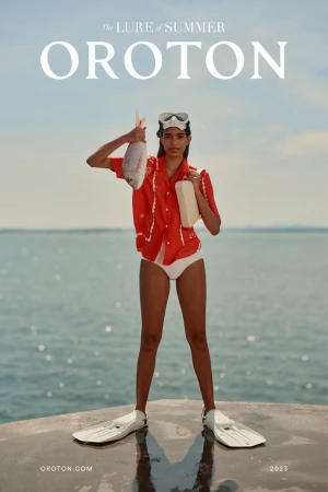 A dark-skinned woman in swim fins poses outdoors with a fish and boxy purse against a blue sea backdrop for a fashion advertisement.