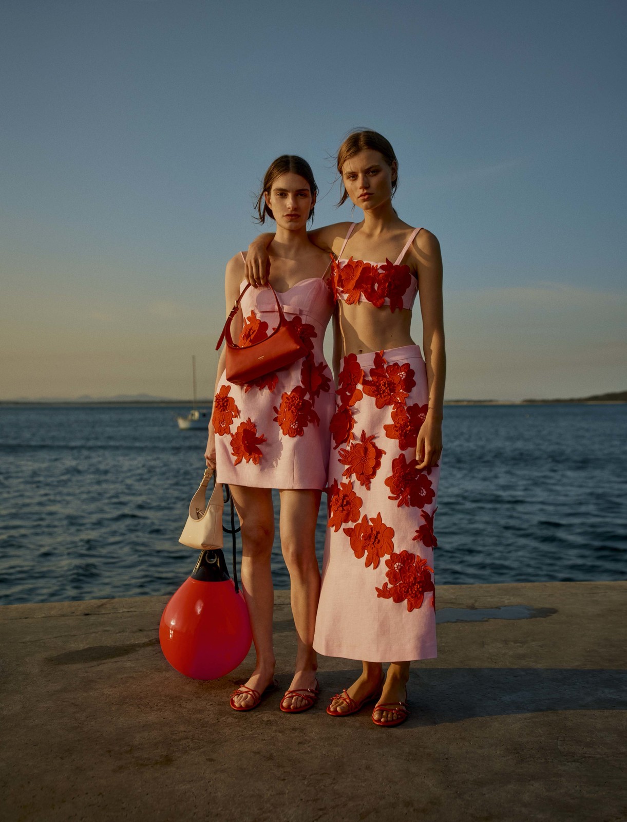 Two women pose outdoors in matching pink floral outfits by the sea. One woman has a purse and the other is holding a red buoy.