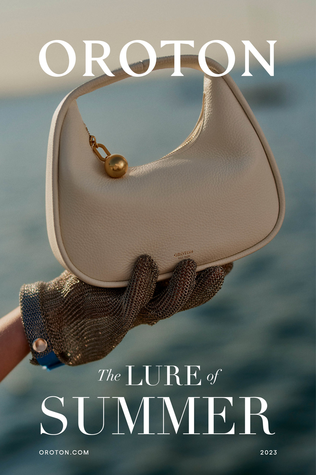 Oroton purse in cream color held up by a hand with metal mesh glove in front of an out-of-focus ocean background, fashion