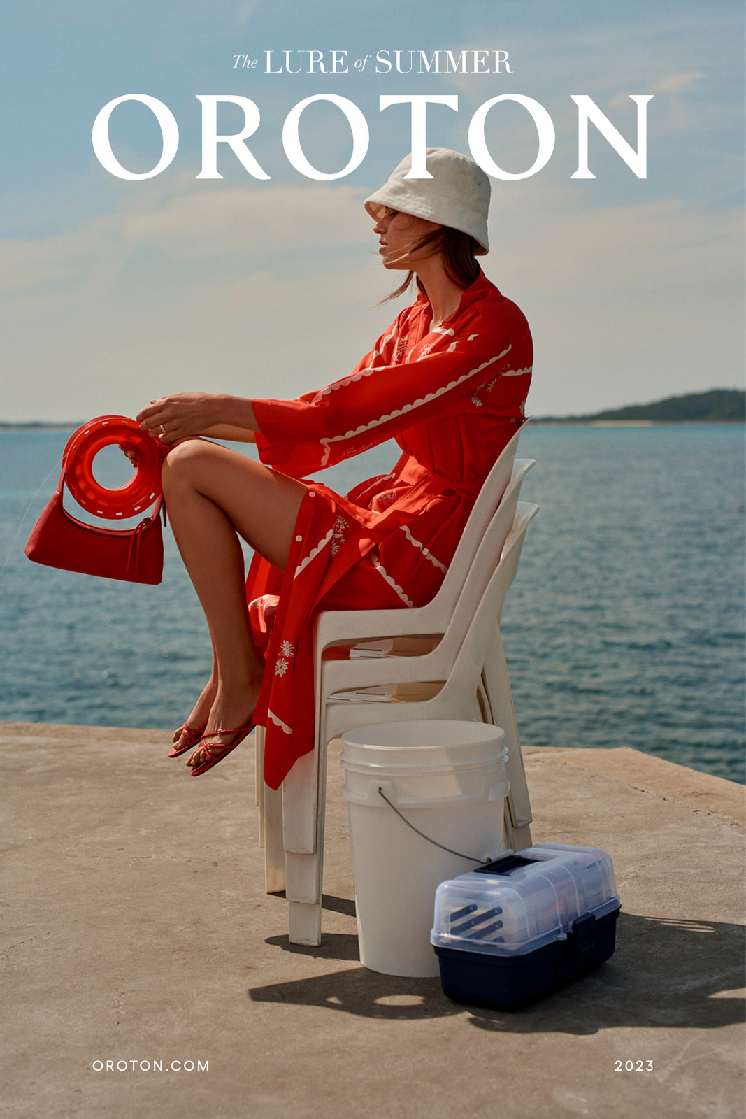 A woman in a red dress and white bucket hat sits on a white plastic chair holding a red handbag, with a white bucket and carrier nearby.