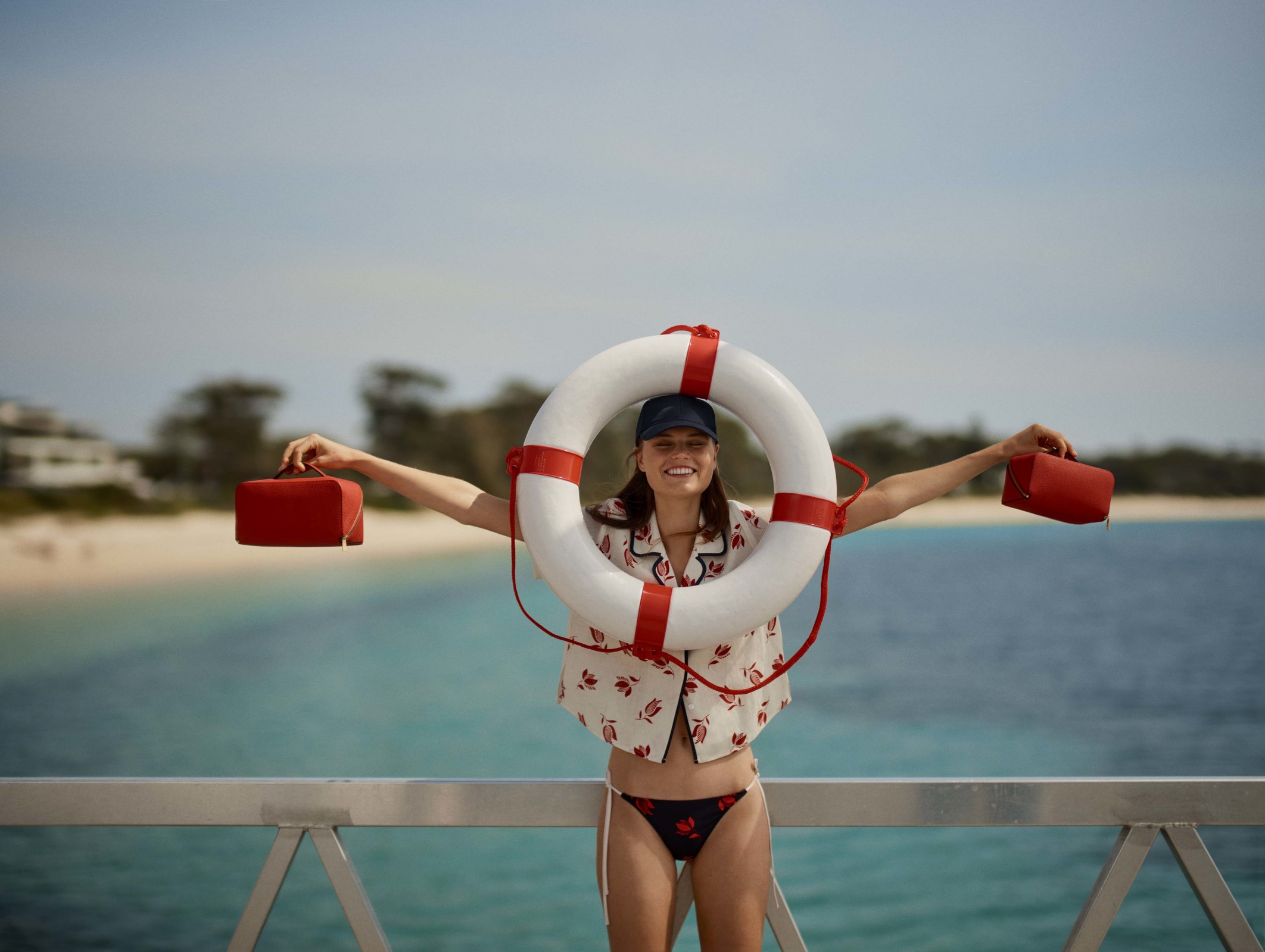 Woman poses with life preserver on pier with red bags, against sea and sky, wearing patterned shirt and bikini bottom, smiling.