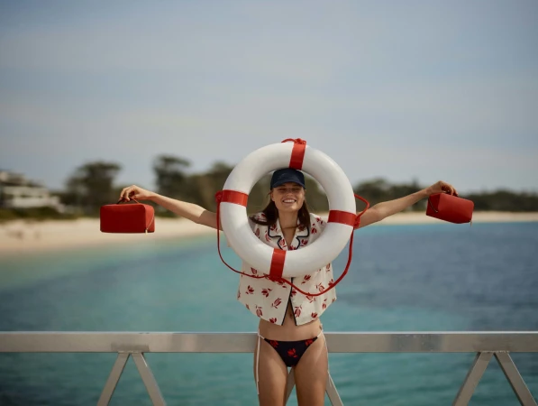 Woman poses with life preserver on pier with red bags, against sea and sky, wearing patterned shirt and bikini bottom, smiling.
