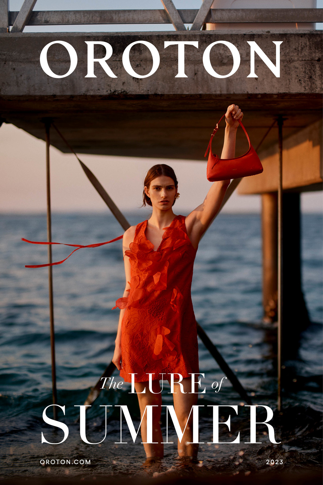 Fashion shot of a woman in a red dress standing in the ocean, holding a red handbag, under a concrete pier, against a sky gradient backdrop.