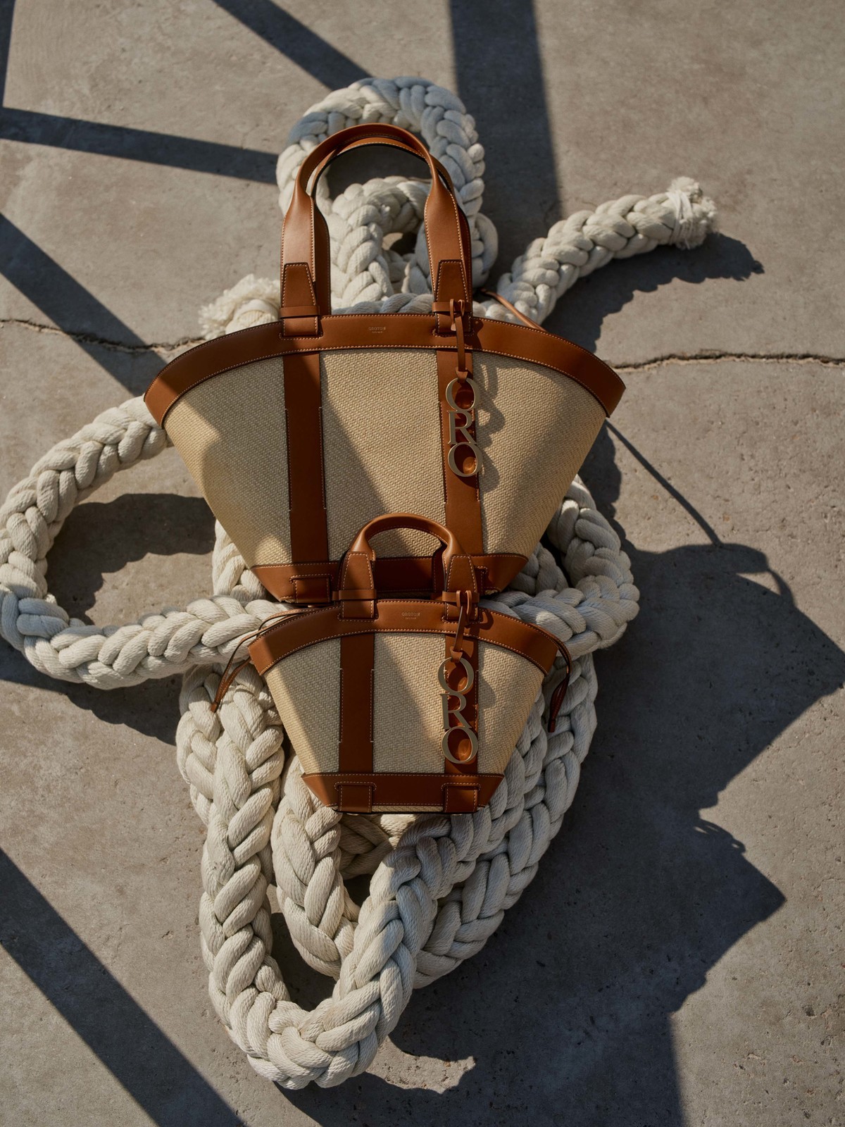Two beige tote bags with brown trim sit atop a thick white rope against a concrete surface, showcasing minimalist design and neutral tones.