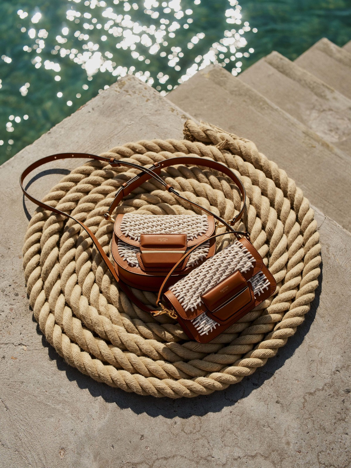Two leather and raffia shoulder bags are displayed on a thick beige rope against a backdrop of sparkling turquoise water.