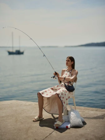 Woman in patterned dress holding a fishing rod while seated on a concrete pier overlooking the ocean