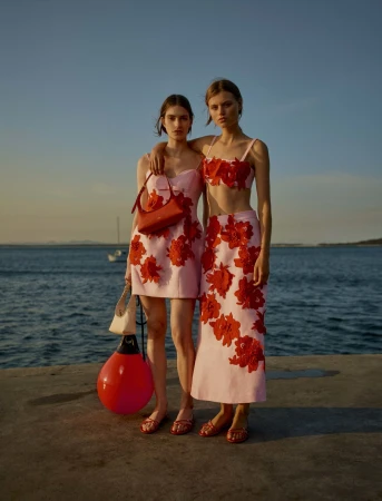 Two female models in pink outfits with red floral applique standing on a pier by the ocean at sunset.