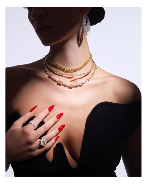 A woman with red nails poses in a studio setting, showcasing three necklaces and multiple rings, complemented by a black top and earrings.