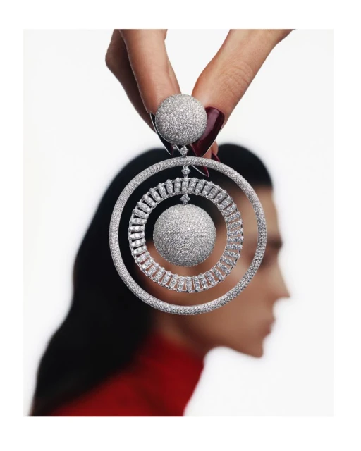 Crystal jewelry earring in a spiral shape held in a woman's hand with a blurred woman's face in the background against a white background.