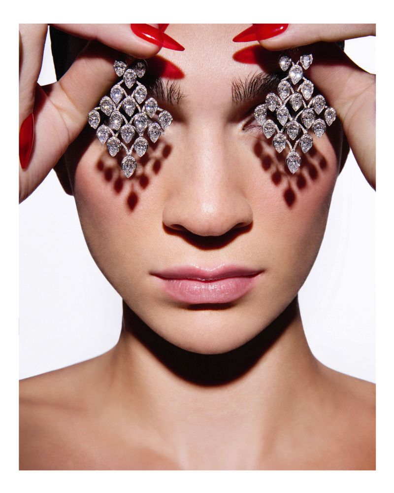 Close-up portrait of a woman with red nails holding large diamond earrings in front of her eyes against a white background.