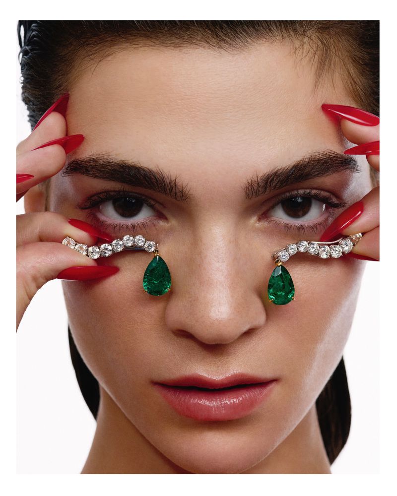 Close-up studio shot of a woman with red fingernails holding jeweled decorations near her eyes featuring diamonds and emeralds.