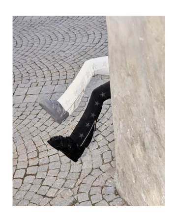Legs of two people wearing star-patterned pants, one black and one off-white, extend from behind a concrete pillar onto a cobblestone surface.