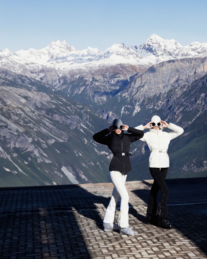 Two women in winter outfits pose on a rooftop with snowy mountains in the background, both wearing hats and sunglasses, looking up.