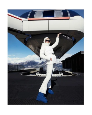 Fashion shot of an adult female model in a white ski suit with blue boots, posing in front of a mountain-top observation deck and snowy mountains.