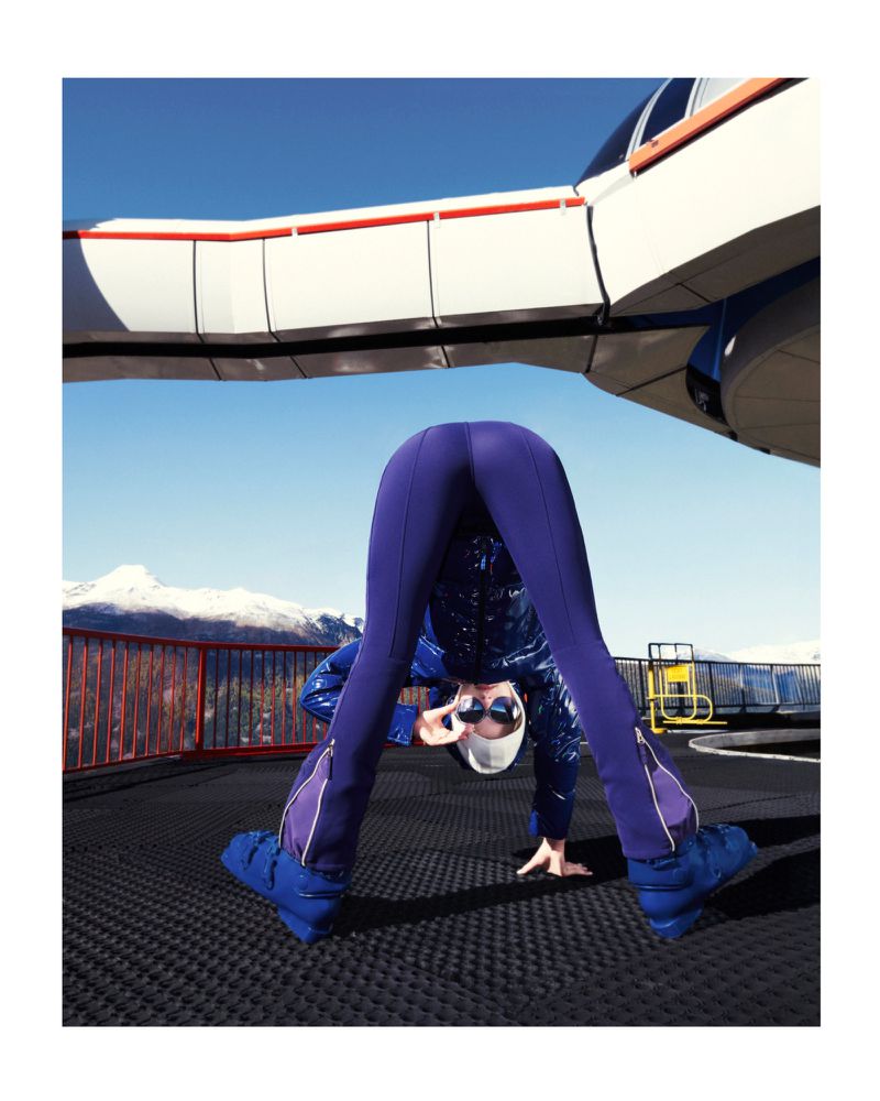 Woman in a headstand with goggles and puffy boots on a rooftop with mountains and a white building in the background.