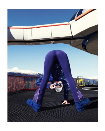 Woman in a headstand with goggles and puffy boots on a rooftop with mountains and a white building in the background.
