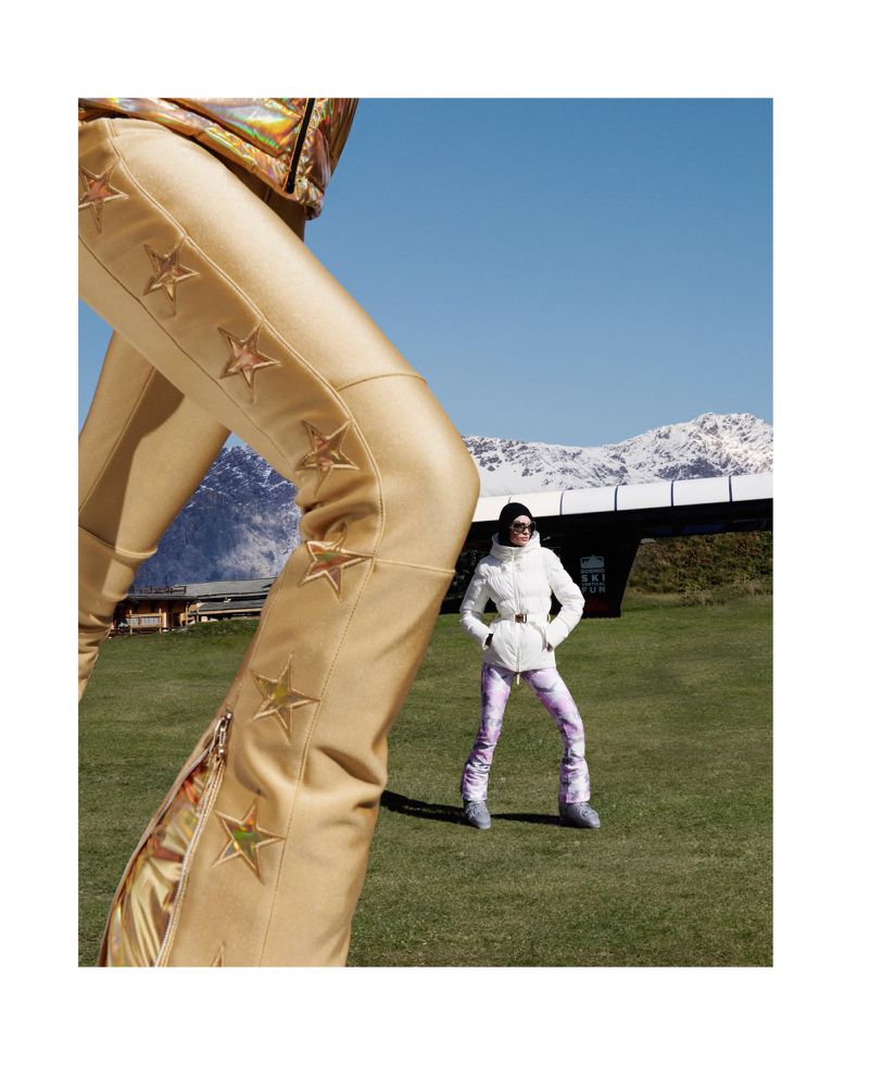 A woman in a white ski suit poses in a grassy field in front of snowy mountains, next to an oversized gold pant leg with star designs.