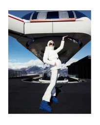 A model in a white puffer jacket and blue boots poses under a large cable car with snow-capped mountains in the background.