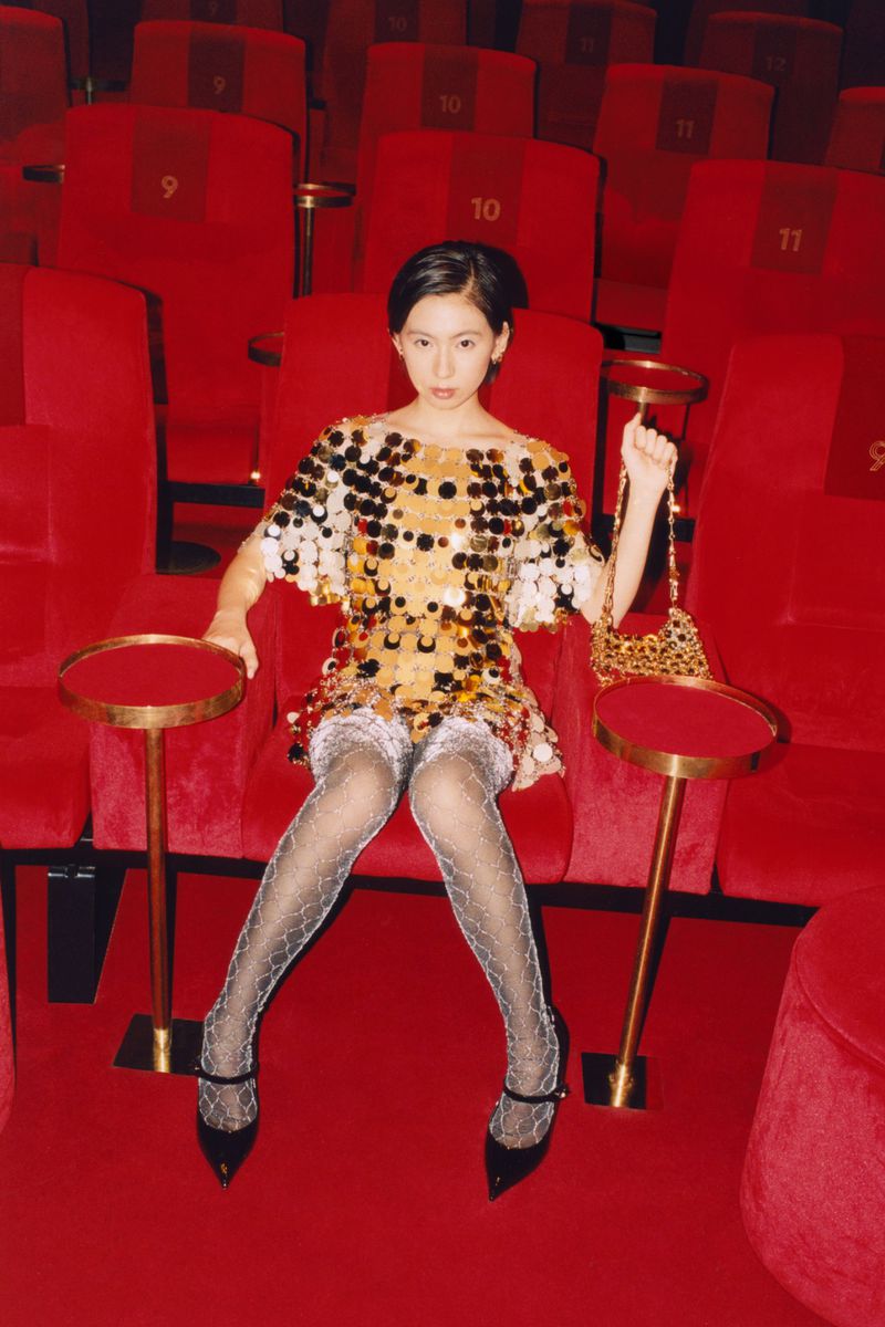 An Asian woman with short dark hair poses in a sequined gold dress with black shoes in a red theatre, holding a leopard print purse.