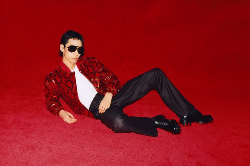 Adult male in patterned jacket and sunglasses lies on red surface, indoors, full body shot, eye-level angle, fashion editorial style.