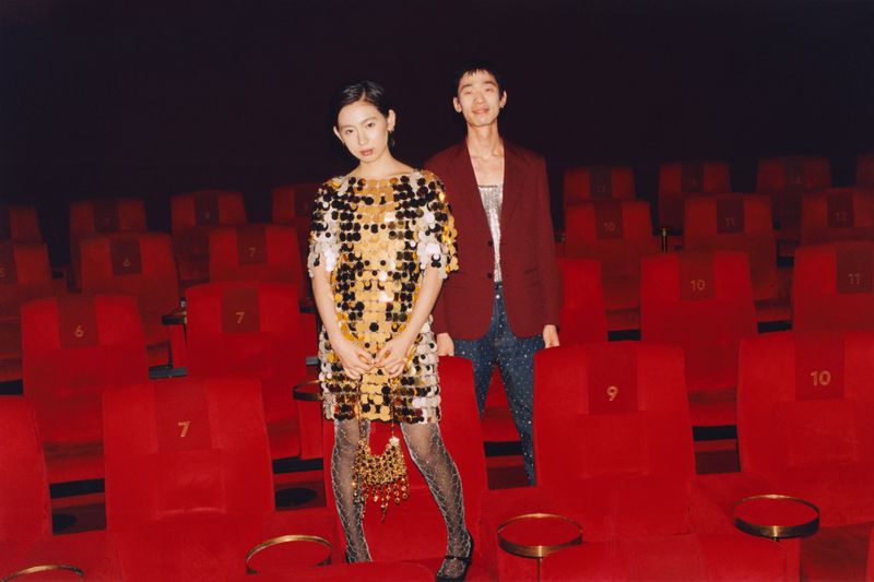 An Asian woman in a gold dress and a man in a maroon jacket pose in a movie theatre with empty red seats, captured in a medium shot.