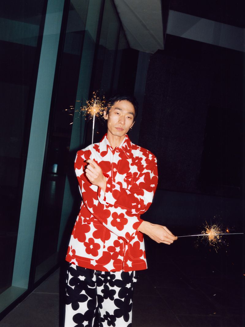 Man in floral print shirt and pants holds sparklers at night near modern building, creating a festive and stylish visual moment.