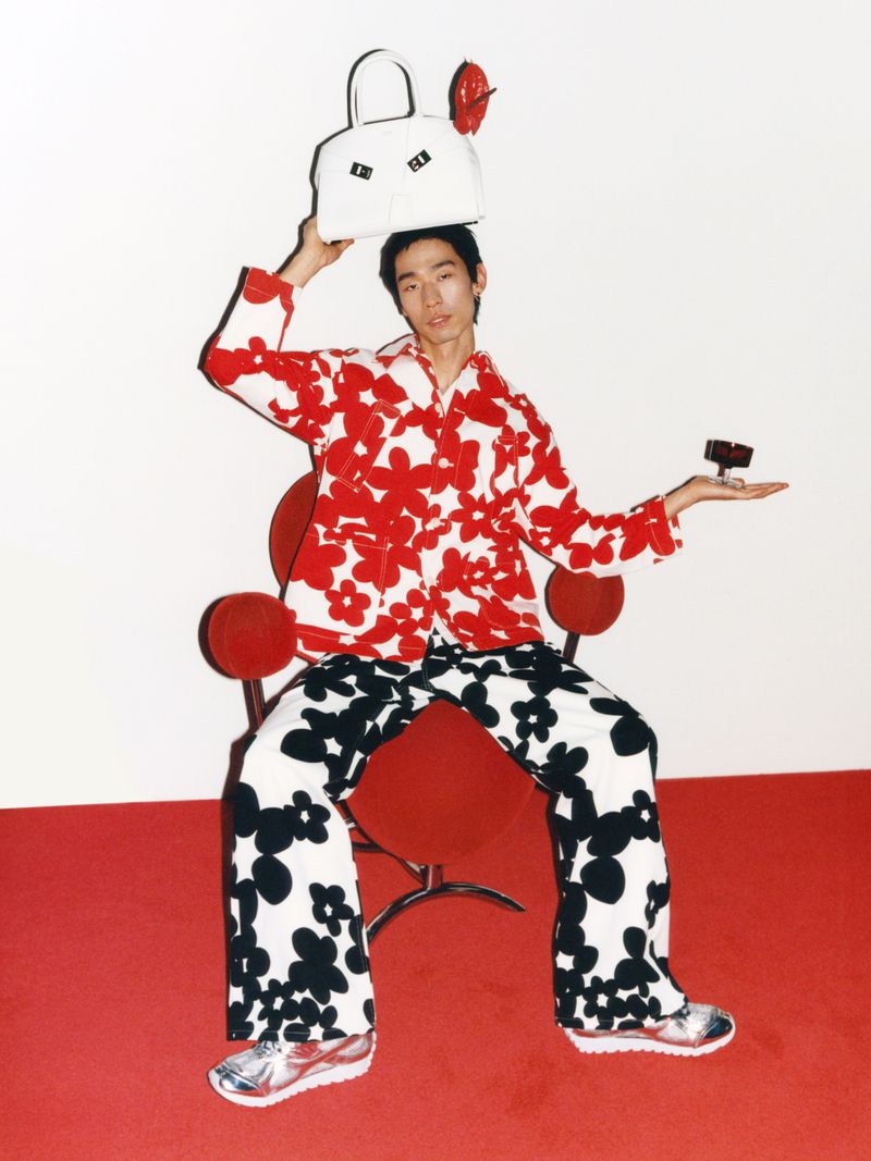 Asian adult sits on a red chair holding a white bag over his head and a small box in his hand, wearing floral patterns.