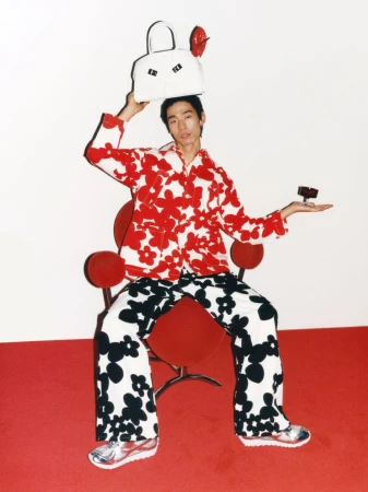 Asian adult sits on a red chair holding a white bag over his head and a small box in his hand, wearing floral patterns.