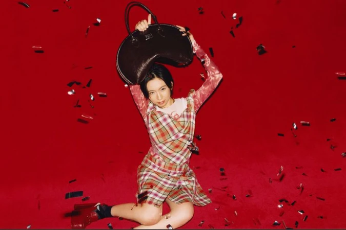 Model seated on a bright red studio floor holding a large dark handbag overhead amidst falling confetti.