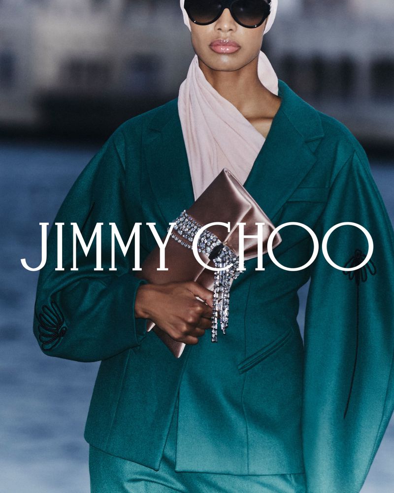 Jimmy Choo campaign featuring an adult female model in a teal suit holding a crystal-embellished clutch bag against a blurred background.
