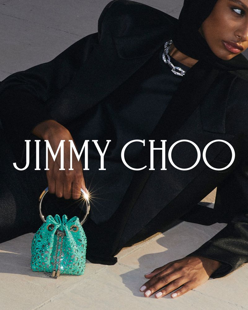 An adult woman wearing a black outfit poses in a studio with a shiny silver necklace and a teal Jimmy Choo handbag with jewel embellishments.