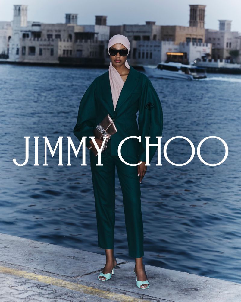 An adult female fashion model in a green suit and headscarf poses with a handbag against an urban waterscape backdrop for Jimmy Choo.