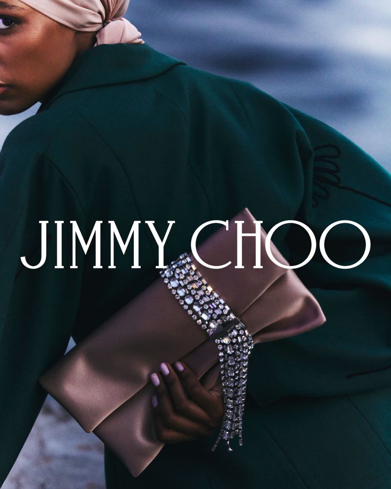 A dark-skinned model wears a green jacket and headscarf while holding a Jimmy Choo clutch with crystal detailing, set against an outdoor backdrop.
