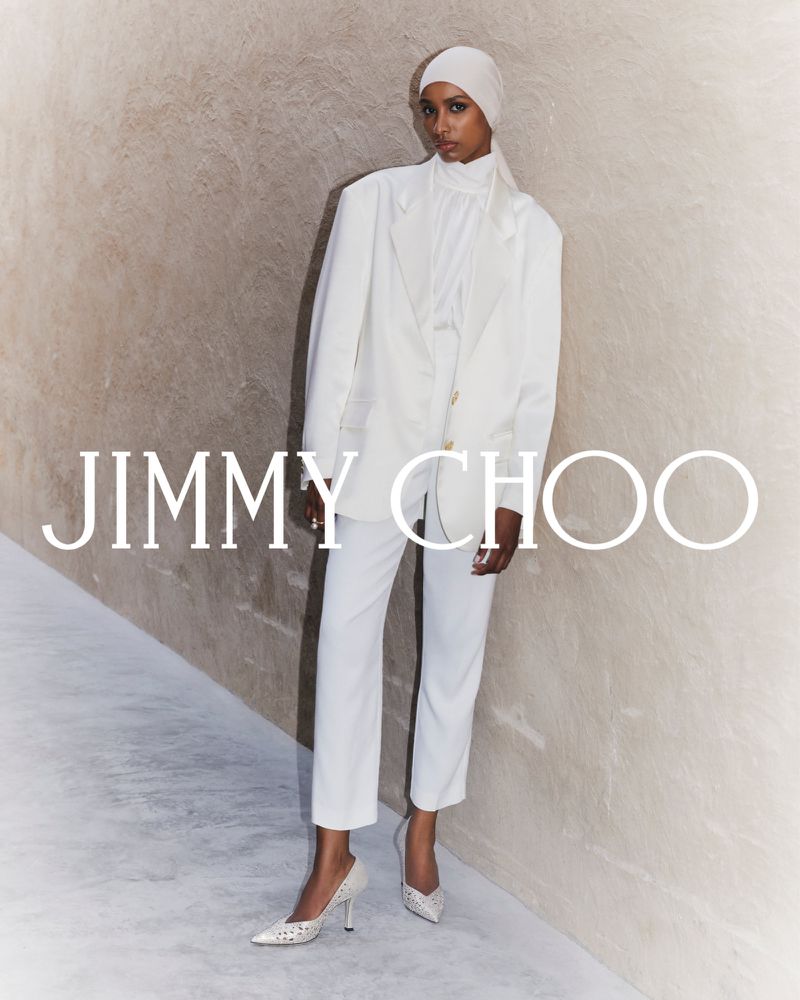 A woman in a white pantsuit and headscarf stands in front of a textured wall, wearing pointed-toe heels, with the JIMMY CHOO logo.