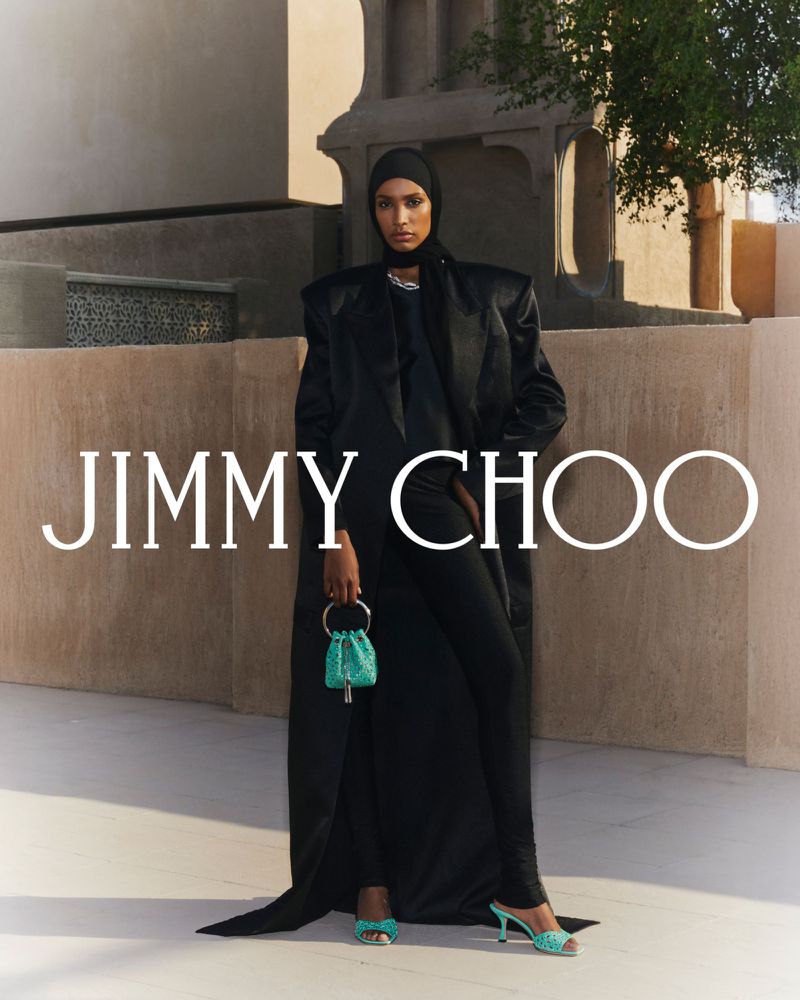 A woman in a black outfit poses with a turquoise purse and matching heels against a neutral backdrop for Jimmy Choo fashion.
