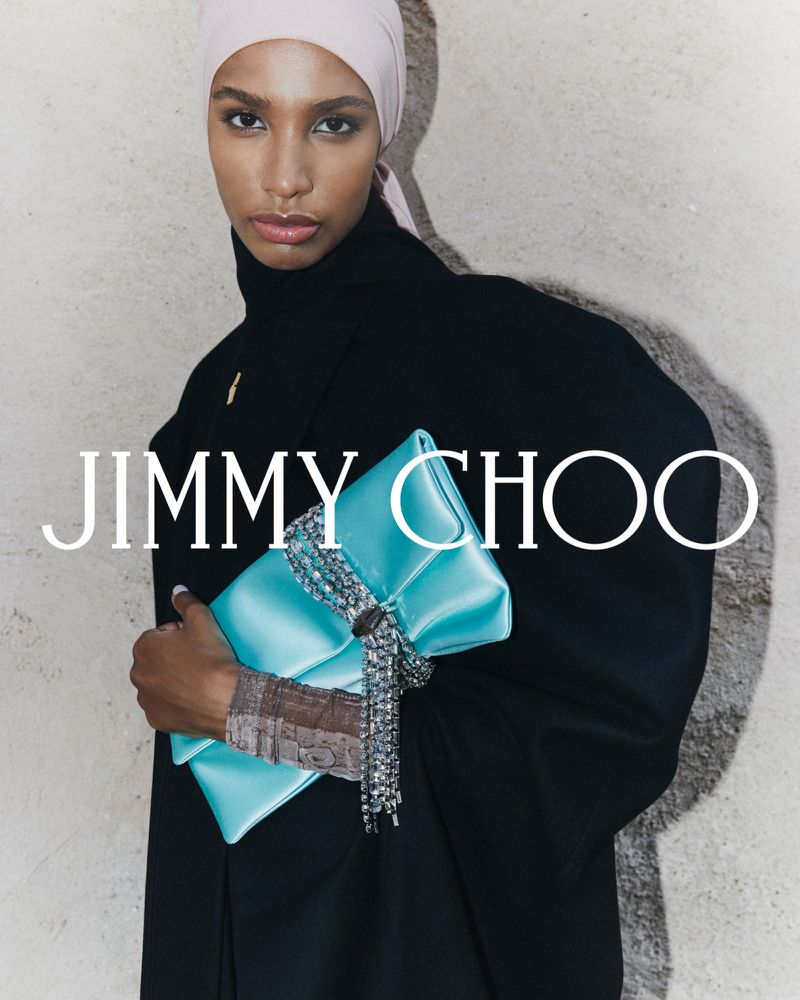Fashion shot of a woman in a black top and light pink headscarf holding a blue Jimmy Choo clutch with silver fringe against a textured wall.