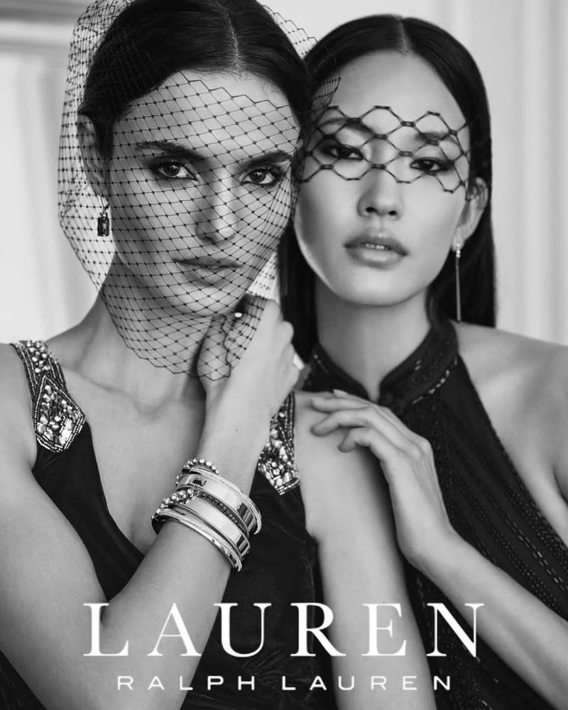 Black and white close-up of two women with veils, adorned dresses, and jewelry, featuring the RALPH LAUREN logo for fashion advertising.