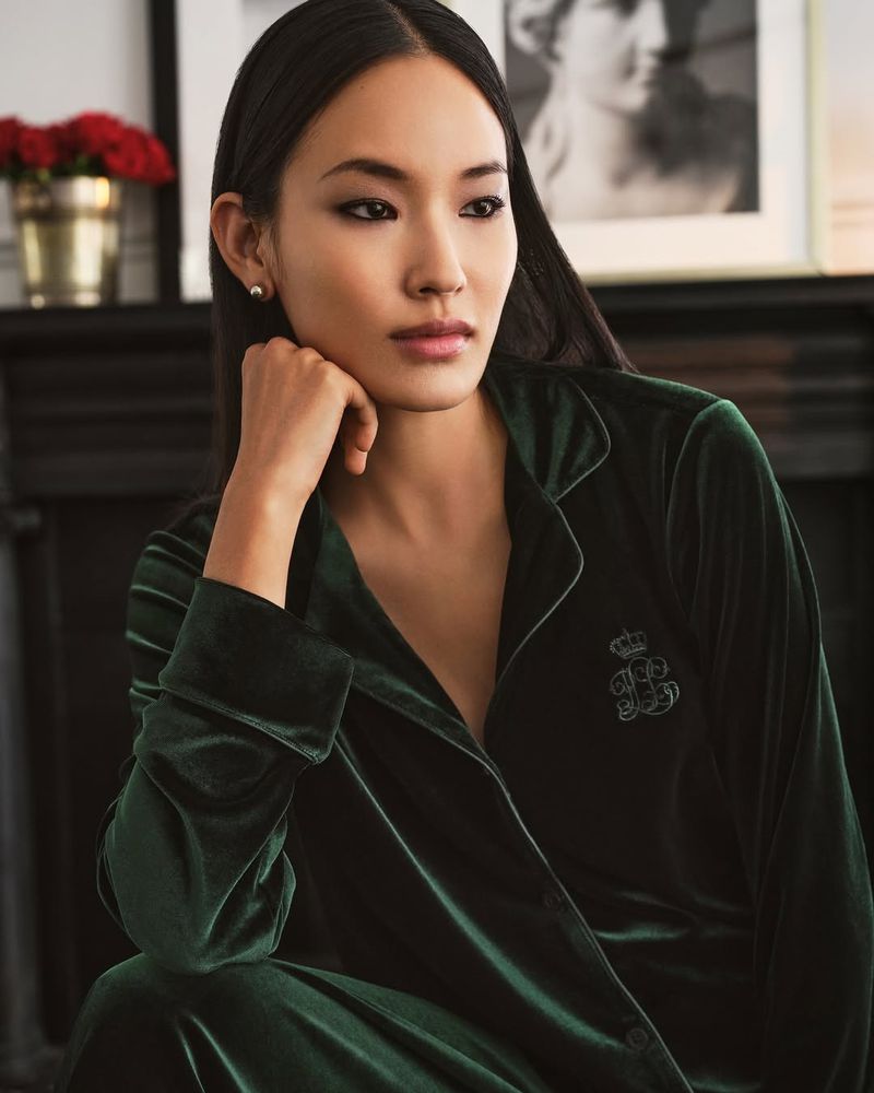 Asian woman in a relaxed pose wearing a dark green velvet pajama set, soft lighting, with floral and framed art in the background.